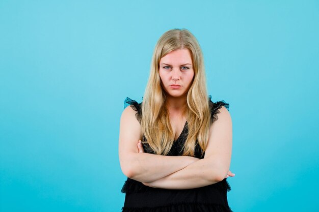 Ernsthaft blondes Mädchen schaut in die Kamera, indem sie die Arme auf blauem Hintergrund verschränkt