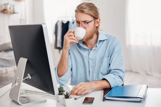 Ernsthaft konzentriert auf Arbeit Büroangestellte mit hellem Haar, Bart in lässigem Outfit und Brille, bereitet Bericht vor, benutzt Tastatur, trinkt Kaffee, arbeitet während der Mittagspause, sitzt gegen Büroeinrichtung.