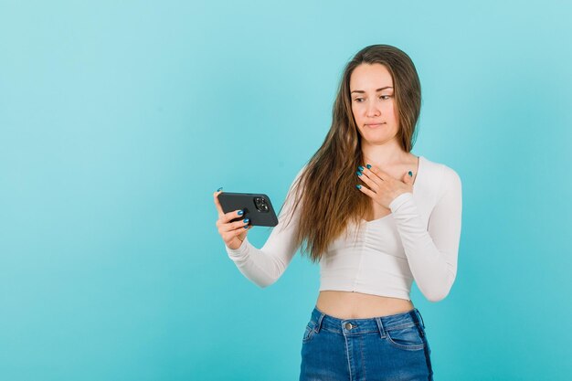 Ernsthaft, Mädchen schaut auf den Smartphone-Bildschirm, indem sie die Hand auf der Brust auf blauem Hintergrund hält