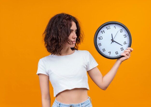 Ernsthaft schauende junge schöne Frau, die Uhr hält und sie auf isolierter orange Wand mit Kopienraum betrachtet
