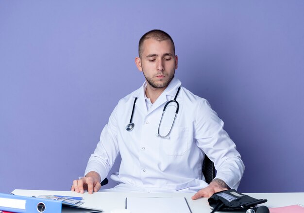 Ernsthaft schauender junger männlicher Arzt, der medizinische Robe und Stethoskop trägt, sitzt am Schreibtisch mit Arbeitswerkzeugen, die Hände auf Schreibtisch setzen und auf Schreibtisch lokalisiert auf lila Wand schauen