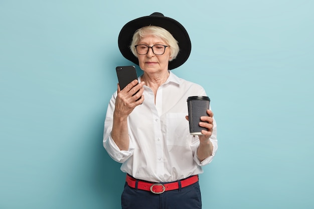 Ernsthafte ältere Frau konzentriert sich auf Smartphone-Gerät, sucht Informationen im Internet, liest Artikel online, trinkt Kaffee zum Mitnehmen, verwendet einfache Anwendung für Rentner, trägt stilvolles Outfit, posiert drinnen