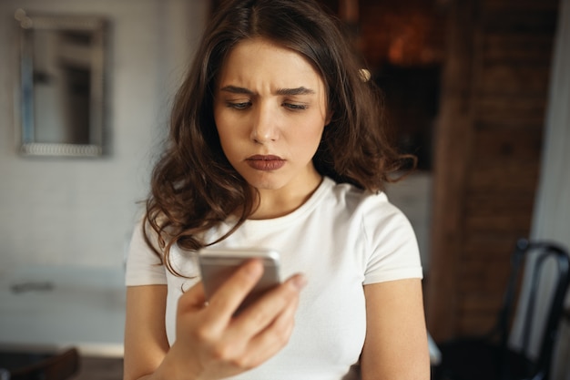 Ernsthafte attraktive junge Frau, die Smartphone hält und die Stirn runzelt und traurige Nachrichten liest