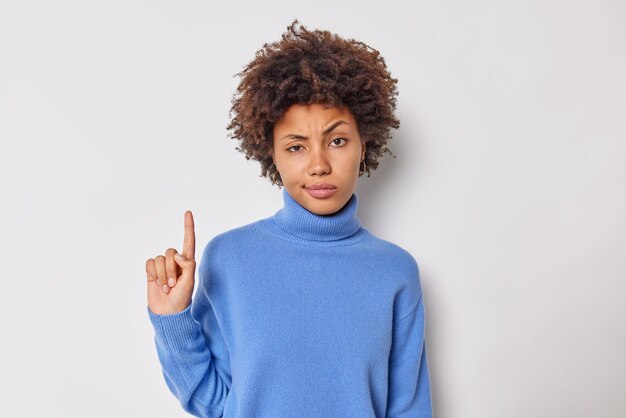 Ernsthafte aufmerksame strenge Frau zeigt mit dem Zeigefinger oben zeigt etwas über Kopf hat einen selbstbewussten Gesichtsausdruck trägt einen lässigen blauen Pullover einzeln auf weißem Hintergrund. Schau dir das einfach an