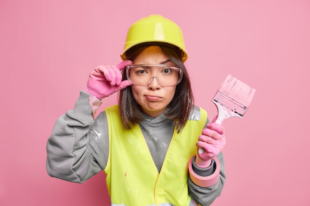 Ernsthafte aufmerksame Wartungsarbeiterin hält Hand an transparenter Schutzbrille, die mit dem Umbau des Hauses beschäftigt ist, hält Malpinsel trägt Schutzhelmuniform