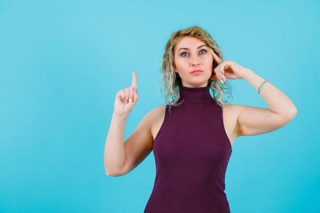 Ernsthafte blonde Frau denkt, dass sie eine Hand an der Schläfe hält und eine Minute mit der anderen auf blauem Hintergrund zeigt