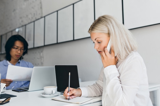 Kostenloses Foto ernsthafte blonde frau, die am telefon spricht und etwas auf papier schreibt