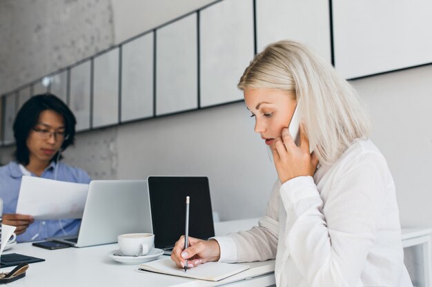 Ernsthafte blonde Frau, die am Telefon spricht und etwas auf Papier schreibt