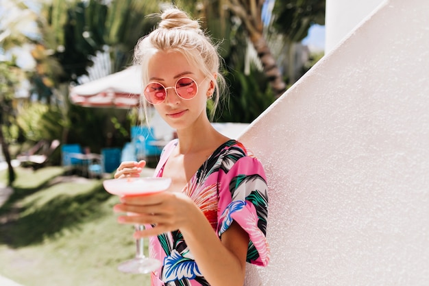 Ernsthafte blonde Frau mit sanftem Lächeln, das mit Cocktail aufwirft.