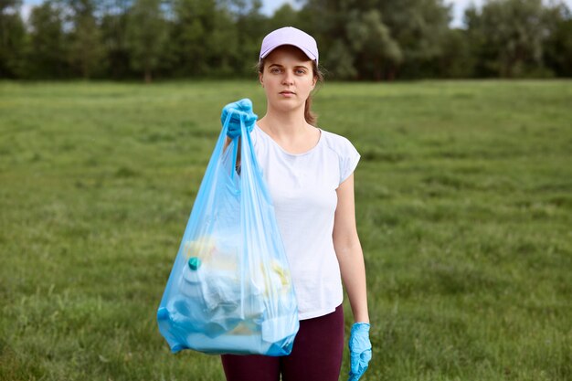 Ernsthafte Dame in der Baseballmütze, die Müllsack voller Müll in den Händen hält und es Leuten zeigt