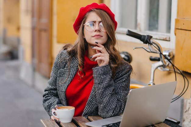 Ernsthafte Frau mit elegantem lockigem Haar, das zur Kamera schaut, während mit Computer im Straßencafé arbeitet