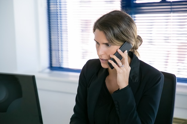 Ernsthafte Geschäftsfrau in der Jacke, die auf Mobiltelefon spricht, während Computer am Arbeitsplatz im Büro verwendet wird. Mittlerer Schuss. Digitales Kommunikations- und Multitasking-Konzept