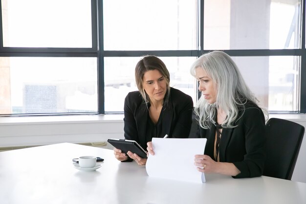 Ernsthafte Geschäftsfrauen diskutieren Berichte. Zwei weibliche Fachleute sitzen zusammen, halten Dokumente, verwenden Tablette und sprechen. Kommunikationskonzept
