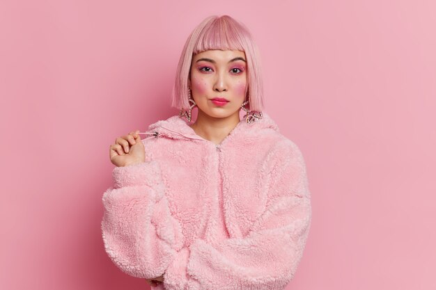Ernsthafte hübsche asiatische Frau mit trendigem rosa Haar im Wintermantel gekleidet hat helle lebendige Make-up-Posen