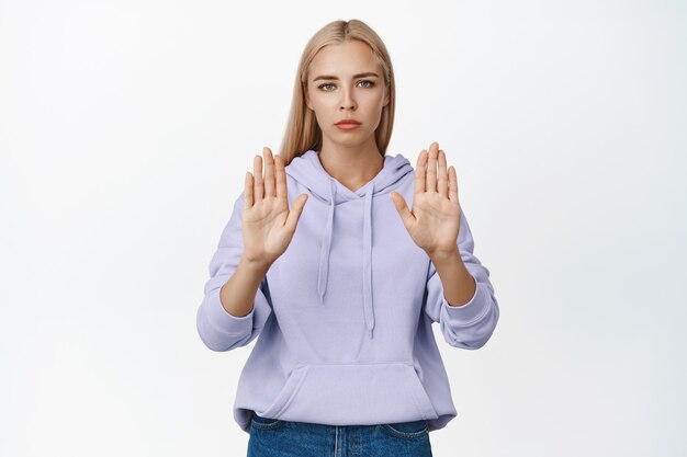 Ernsthafte junge blonde Frau stößt Sie zurück, zeigt Halt, keine Geste, lehnt etwas ab, steht auf Weiß.
