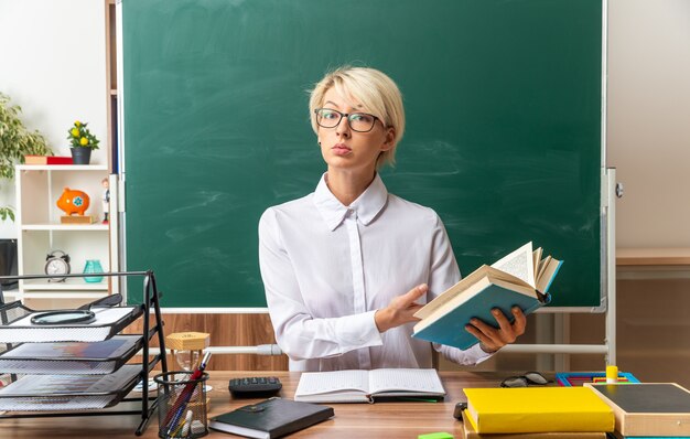 Ernsthafte junge blonde Lehrerin mit Brille, die am Schreibtisch mit Schulmaterial im Klassenzimmer sitzt und auf offenes Buch mit Blick auf die Vorderseite zeigt