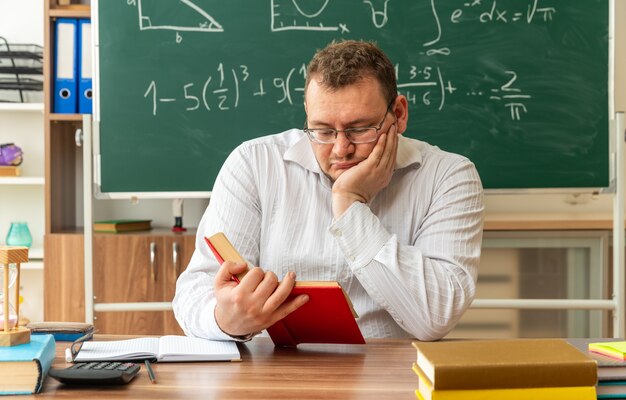 Ernsthafte junge blonde Lehrerin mit Brille, die am Schreibtisch mit Schulwerkzeugen im Klassenzimmer sitzt und ein Buch liest, das die Hand auf dem Gesicht hält