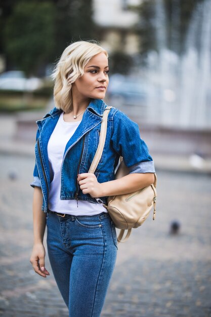 Ernsthafte junge blonde Mädchenfrau auf Straßenwegquadrat-Fontain, gekleidet in Blue Jeans Suite mit Tasche auf ihrer Schulter in sonnigem Tag