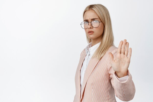 Ernsthafte junge Chefin, die nein sagt, die Geste des Stoppverbots zeigt, in Anzug und Brille auf Weiß steht.