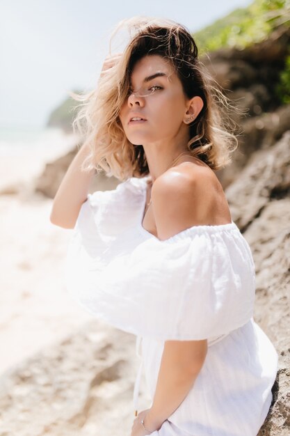 Ernsthafte junge Dame, die während der Ruhe auf der Insel schaut. Außenfoto der prächtigen Frau im weißen Kleid, das auf Natur aufwirft.