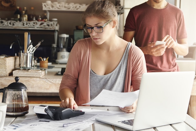 Ernsthafte junge Frau, die rechteckige Brille trägt, die Ausgaben berechnet, während Familienbudget mit generischem Laptop und Taschenrechner zu Hause tut