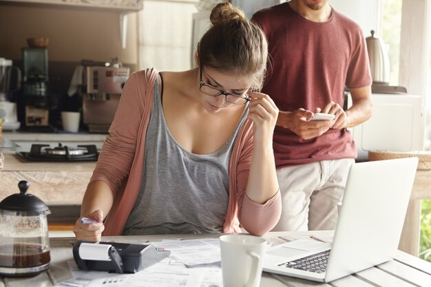 Ernsthafte junge Frau mit Haarknoten, die Brillen tragen, die durch Finanzen schauen