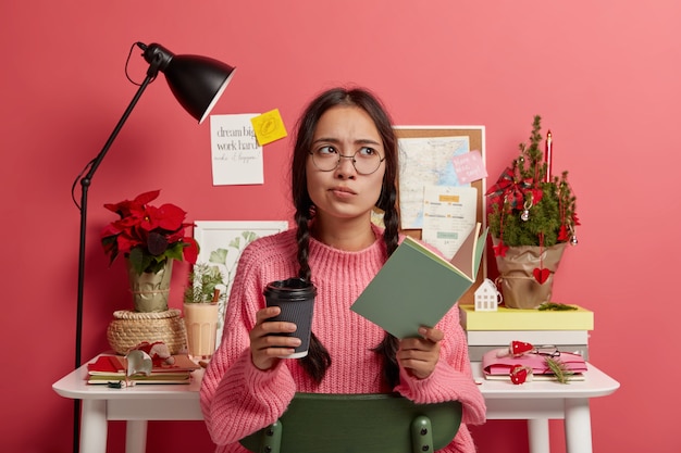 Ernsthafte kontemplative Frau trägt Brille und übergroßen Pullover, hält Pappbecher Kaffee, Lehrbuch für Bildung