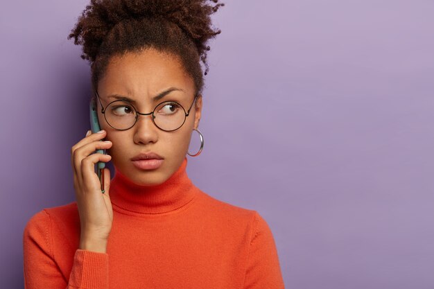 Ernsthafte lockige Frau hat gesunde Haut, hält modernes Handy, trägt Brille und lässigen Rollkragenpullover, isoliert auf violetter Wand