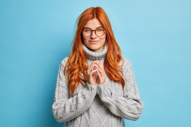 Ernsthafte rothaarige junge europäische Frau entwirft etwas steepls Finger und sieht mit der Absicht zu tun, trägt optische Brille grauen Strickpullover.