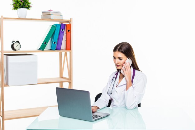 Ernsthafte Ärztin, die an ihrem Schreibtisch sitzt, während sie jemanden über das Telefon anruft und ihren Laptop benutzt