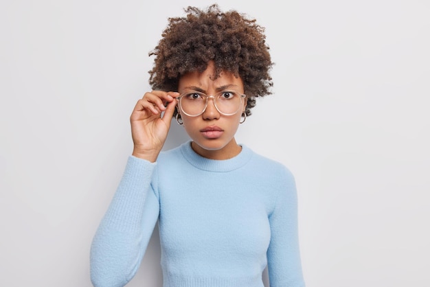 Kostenloses Foto ernsthafte schöne frau mit dunklem lockigem haar blickt aufmerksam durch eine transparente brille in lässigen blauen pullover isoliert auf weißem hintergrund in die kamera und sieht aufmerksam etwas an