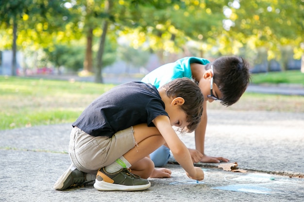 Ernsthafte schwarzhaarige Jungen, die mit bunten Kreiden sitzen und zeichnen. Seitenansicht. Konzept für Kindheit und Kreativität