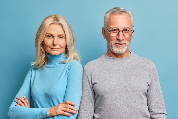 Ernsthafte selbstbewusste blonde hübsche Frau steht mit verschränkten Armen in der Nähe ihres Mannes Pose für das gemeinsame Foto gekleidet in lässigen Rollkragenpullover isoliert über blaue Wand