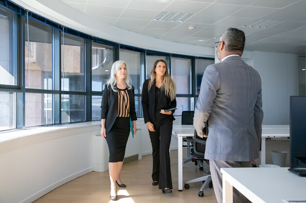 Ernsthafte selbstbewusste Geschäftsfrauen, die zu Mann im Anzug im Büroinnenraum gehen. Volle Länge, Rückansicht. Geschäftstreffen-Konzept