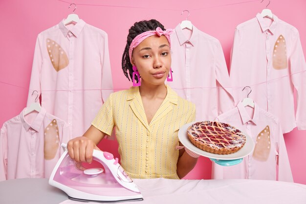 Ernsthafte selbstbewusste Hausfrau trägt Stirnband Hauskleidung Bügeleisen Wäsche backt köstliche Kuchen Posen im Morgenmantel. Housekeeping-Prozess