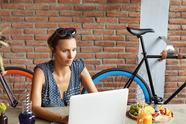 Ernsthafte und selbstbewusste selbständige Frau, die lässig gekleidet ist und auf einem modernen Laptop-Computer tippt, während sie ein elektronisches Gerät für Fernarbeit verwendet