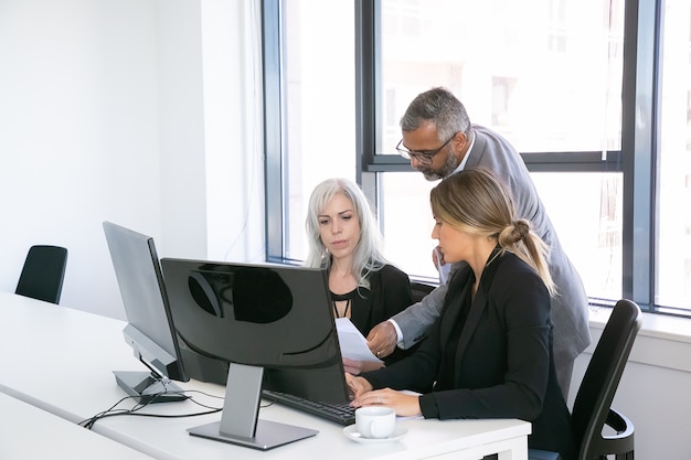 Ernsthafte Unternehmensgruppe von drei, die Berichte analysiert, mit Monitoren am Arbeitsplatz sitzt, Papiere hält, überprüft und diskutiert. Speicherplatz kopieren. Geschäftstreffen-Konzept