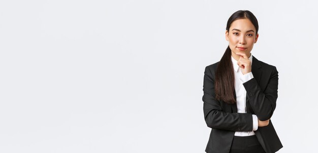 Ernsthafte zufriedene asiatische Geschäftsfrau hat eine interessante Idee, berührt das Kinn und schaut schlau in die Kamera, steht nachdenklich und denkt, während sie im Anzug auf weißem Hintergrund steht