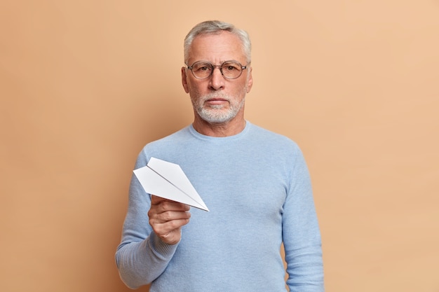 Ernsthafter älterer Mann mit dickem Bart hält Papierflugzeug schaut selbstbewusst vorne hält Papierflugzeug trägt optische Brille lässig Pullover posiert über beige Wand