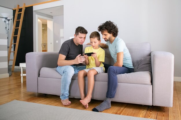Ernsthafter Junge, der Online-Spiel auf Handy spielt, seine zwei Väter sitzen in seiner Nähe und schauen auf den Bildschirm. Familie zu Hause und Kommunikationskonzept