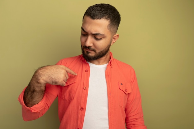 Ernsthafter junger bärtiger Mann im orangefarbenen Hemd, der mit dem Finger auf sich zeigt, der über der hellen Wand steht