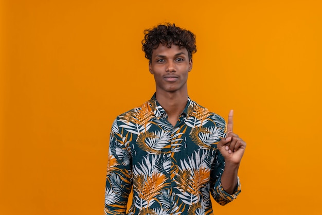 Ernsthafter junger gutaussehender dunkelhäutiger Mann mit lockigem Haar im bedruckten Hemd der Blätter, das eine Geste mit Zeigefinger auf einem orangefarbenen Hintergrund macht