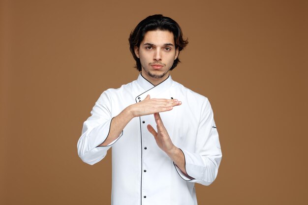 ernsthafter junger männlicher Koch in Uniform, der in die Kamera schaut und eine Timeout-Geste zeigt, die auf braunem Hintergrund isoliert ist