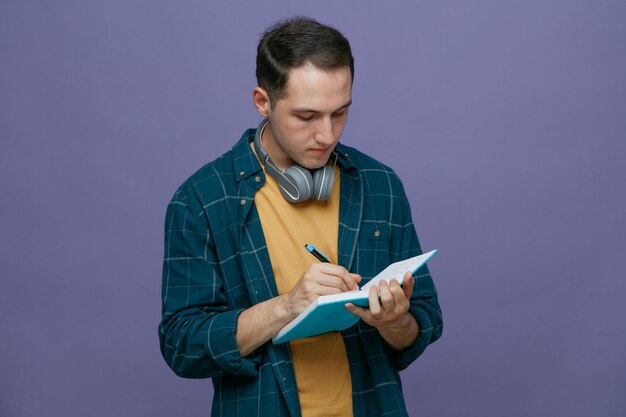 Ernsthafter junger männlicher Student, der Kopfhörer um den Hals trägt und mit Stift auf einem Notizbuch schreibt, das auf violettem Hintergrund isoliert ist