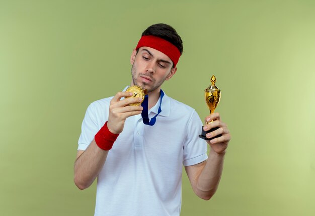 ernsthafter junger sportlicher Mann im Stirnband mit Goldmedaille um den Hals hält seine Trophäe, die über grüner Wand steht