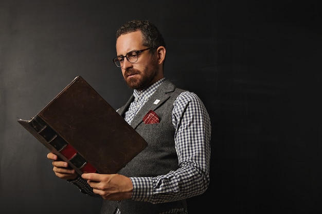 Ernsthafter männlicher Lehrer in Tweedweste und kariertem Hemd, das ein großes altes Buch an einer Tafel liest