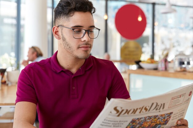 Ernsthafter männlicher Unternehmer beginnt den Tag mit der Morgenzeitung, analysiert Nachrichten in der Presse, trägt eine optische Brille für gute Sicht, trägt ein lässiges T-Shirt und konzentriert sich auf das Lesen von Artikeln in der Cafeteria.