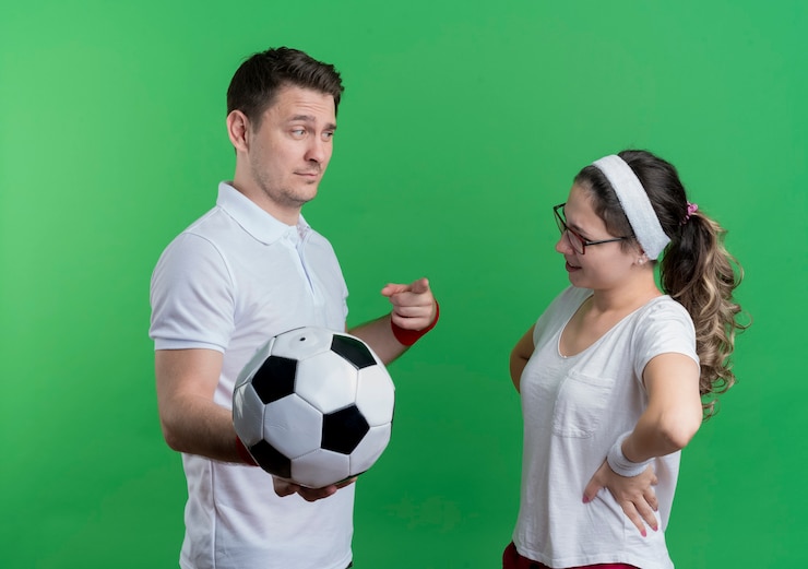 Kostenlose Foto ernsthafter mann des jungen sportlichen paares, der fußball hält, der seine lächelnde freundin über grün betrachtet