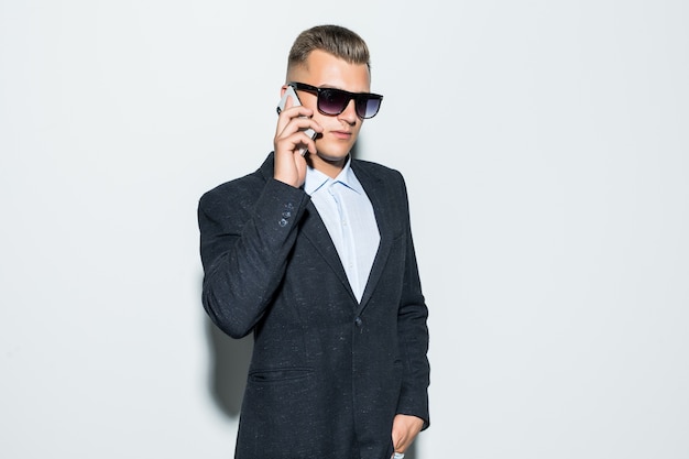 Ernsthafter Mann in Suite und Sonnenbrille spricht auf seinem Handy vor der hellen Wand