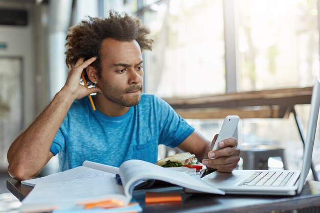 Ernsthafter Mann mit buschigem Haar, der mit wissenschaftlicher Literatur arbeitet und Artikel unter Verwendung moderner Technologien schreibt, um notwendige Informationen in seinem Telefon zu finden